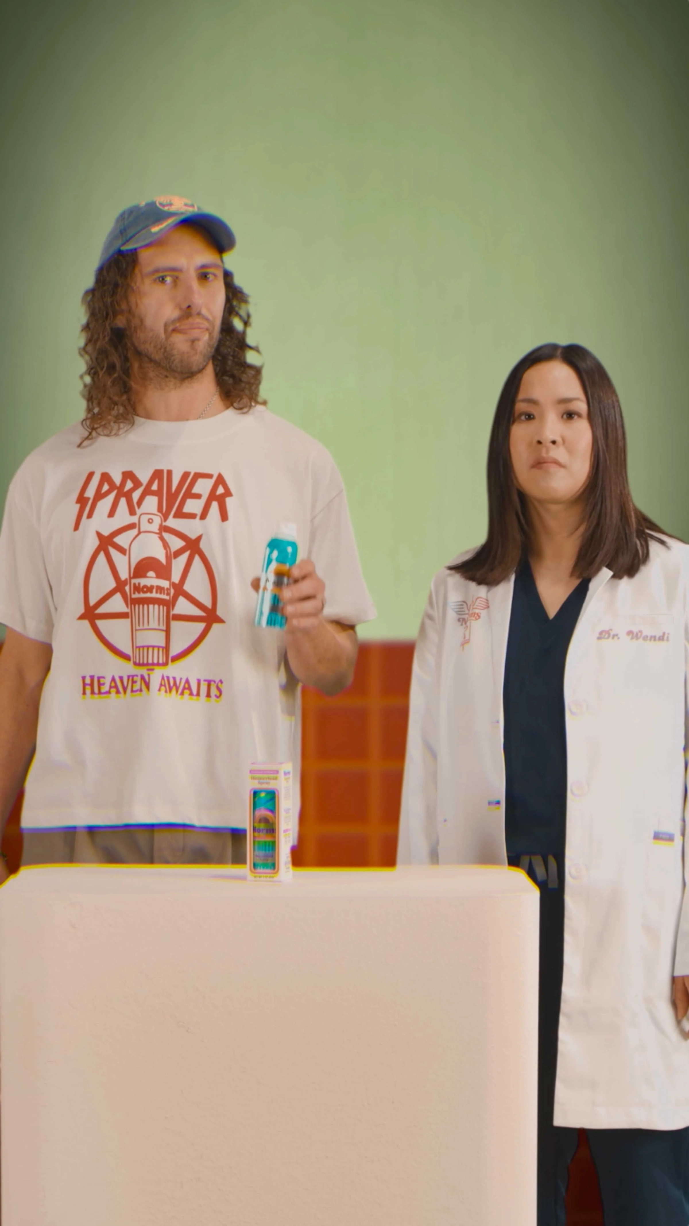 Man in 'Sprayer' t-shirt holding a can, standing next to a woman in a lab coat against a green background. introductory video to norms hemorrhoid relief spray