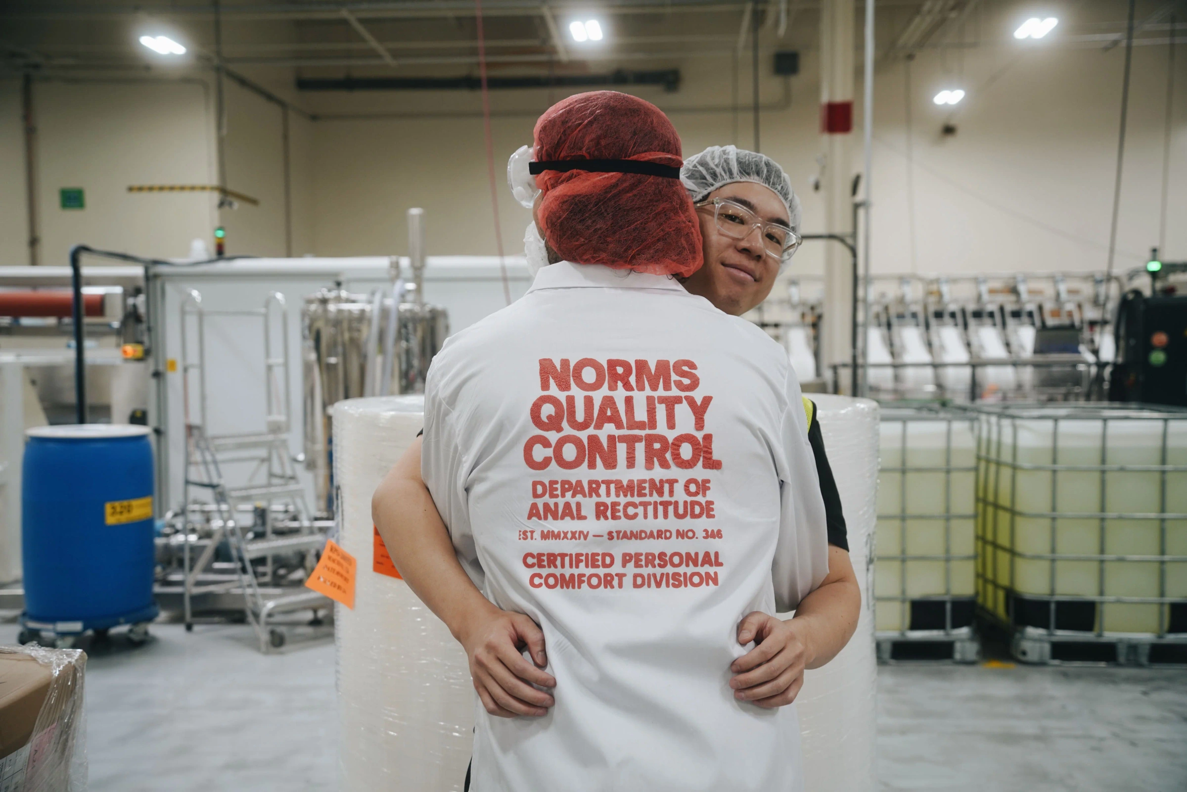 Two people hugging in a factory setting with one wearing a 'Norms Quality Control' shirt.