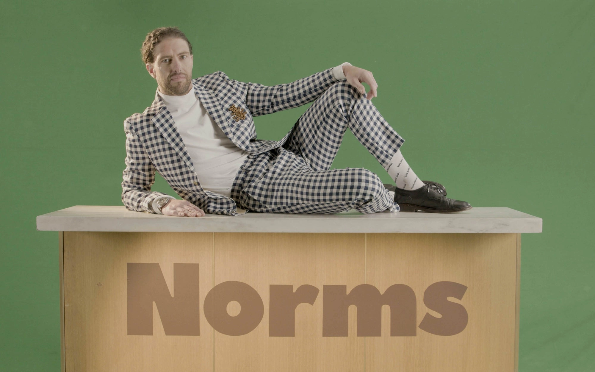 Man in a checkered suit lying on a desk with the Norms logo, green background